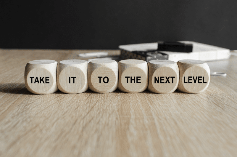 Wooden blocks on a table spell "TAKE IT TO THE NEXT LEVEL." Background includes a blurred book and pens on a wooden surface.